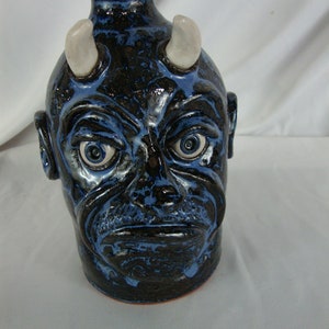 May include: A ceramic jug with a devil face design. The jug is primarily black with blue accents and white horns. The face has detailed features, including eyes, ears, and a mouth. The jug has a rounded body and a narrow neck.