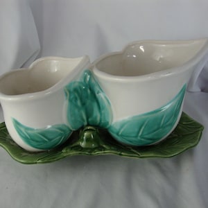 May include: A white ceramic planter with two connected compartments, each with a curved rim. The planter features teal leaf accents and sits on a green leaf-shaped base. A decorative piece for plants.