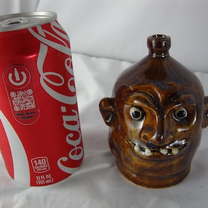 May include: A brown ceramic jug with a face, next to a red Coca-Cola can. The jug has a smiling face with white teeth and black eyes. The can is 12 fl oz (355 mL).