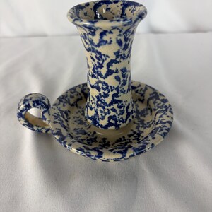 May include: A ceramic candle holder featuring a cream-colored base with a blue speckled sponge-painted pattern. The piece includes a cylindrical candle cup, a circular drip tray, and a small curved handle attached to the side of the base.
