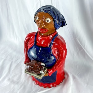 May include: A ceramic figurine of a woman wearing a red dress and blue apron, holding a platter with a brown bird on it.
