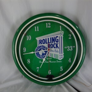 May include: A green circular clock with a white neon ring around the edge. The clock face is green with white numbers and hands. The center of the clock face features a Rolling Rock beer logo with the text "Rolling Rock", "Premium Beer", "Extra Pale", "33", and "Brewed From Mountain Springs".