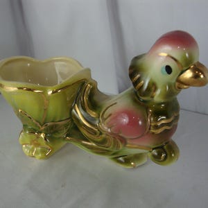May include: A vintage ceramic planter shaped like a bird. The planter features a heart-shaped opening and is painted in shades of green, pink, and gold. The bird has a gold beak and accents, with a small blue eye.
