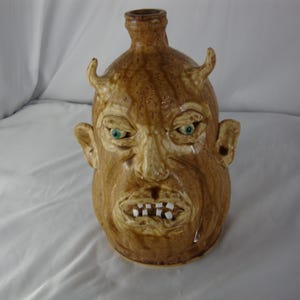 May include: A brown ceramic jug with a face sculpted on the front. The face has green eyes, small horns, and a grimacing mouth with white teeth. The jug has a bottle-like neck.