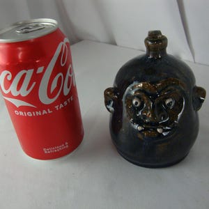 May include: A red Coca-Cola can next to a dark blue ceramic jug with a face design. The jug has a brown face with white eyes and a toothy grin. The can reads "Coca-Cola ORIGINAL TASTE".