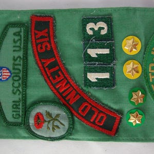 K&ouml;nnte beinhalten: Eine Sammlung von Girl Scout-Abzeichen und Anstecknadeln auf gr&uuml;nem Stoff. Abzeichen sind unter anderem "Girl Scouts USA" und "Old Ninety Six". Gold- und gr&uuml;nfarbene sternf&ouml;rmige Anstecknadeln sind ebenfalls vorhanden. Die Zahl "113" wird ebenfalls angezeigt.