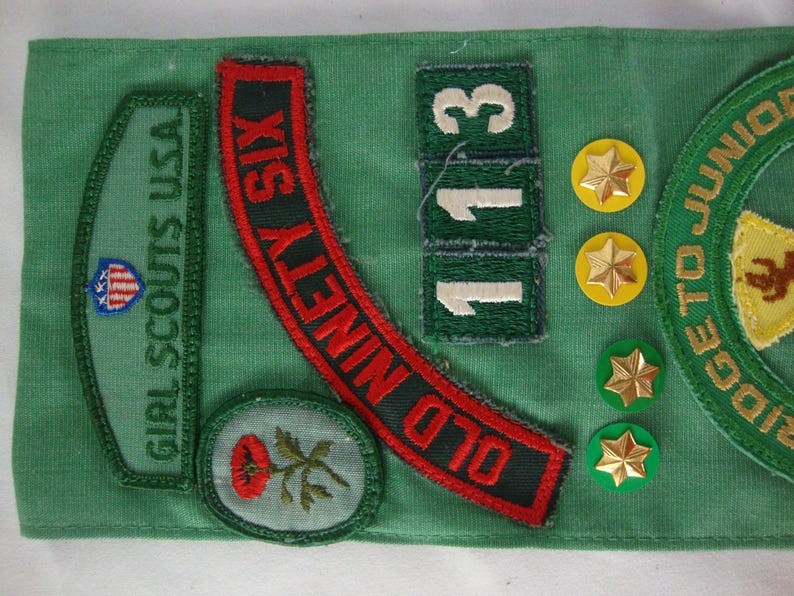 K&ouml;nnte beinhalten: Eine Sammlung von Girl Scout-Abzeichen und Anstecknadeln auf gr&uuml;nem Stoff. Abzeichen sind unter anderem "Girl Scouts USA" und "Old Ninety Six". Gold- und gr&uuml;nfarbene sternf&ouml;rmige Anstecknadeln sind ebenfalls vorhanden. Die Zahl "113" wird ebenfalls angezeigt.