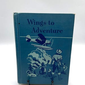 Könnte beinhalten: Ein Vintage-Buch mit dem Titel "Wings to Adventure" mit blauem Einband. Das Cover zeigt ein Doppeldeckerflugzeug, Bäume und Figuren in einer blau-weißen Illustration. Das Buch wird auf einem schwarzen Metallständer präsentiert.