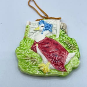 Puede incluir: Adorno de cerámica que representa un pato con un gorro y un chal, de pie cerca de una valla y follaje verde. El pato es blanco con un gorro azul y un chal rojo. El adorno tiene una cuerda dorada para colgar.