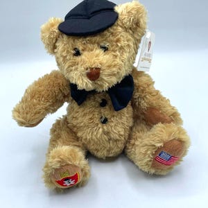 Może przedstawiać: Pluszowy miś w kolorze jasnobrązowym, w czarnej muszce i pasującej czapce. Miś ma brązowy nos i czarne guziczkowe oczy. Na łapie misia wyhaftowana jest amerykańska flaga.