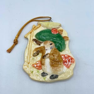 Peut inclure: Ornement en céramique représentant un lapin en veste marron et chapeau vert, tenant un sac. L'ornement a une ficelle dorée pour l'accrocher et comprend des détails de champignons rouges et blancs. Le fond est de couleur beige clair.