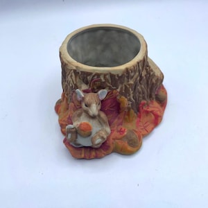 Puede incluir: Una maceta de cerámica con forma de tocón de árbol, con una pequeña figura de animal sentado sosteniendo una nuez. La maceta tiene un exterior marrón y un interior hueco. La base presenta hojas de otoño en tonos naranja y rojo.