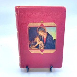 Op de afbeelding: Een rood boek met een goudomrand beeld van een religieuze scène op de omslag. De afbeelding toont een vrouw en een kind. Het boek staat rechtop op een zwarte standaard.