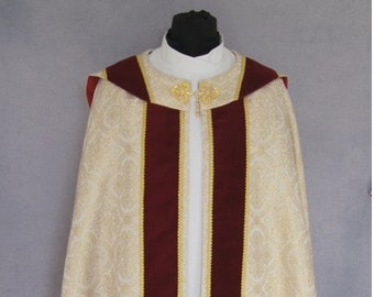 May include: A white liturgical cope with a burgundy velvet trim and gold accents. The cope is worn over a white cassock.