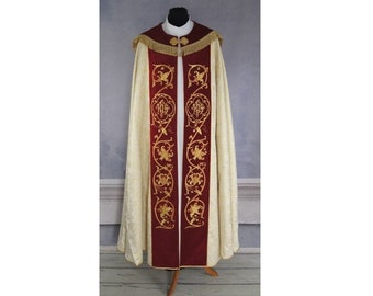 May include: A white and gold brocade cope with a red velvet lining. The cope features gold embroidery with a repeating design of leaves and a monogram.