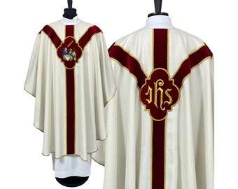 Może przedstawiać: Biała szata liturgiczna z czerwonymi atłasowymi lamówkami i haftowanymi detalami. Przód przedstawia scenę narodzin, a tył monogram "IHS".