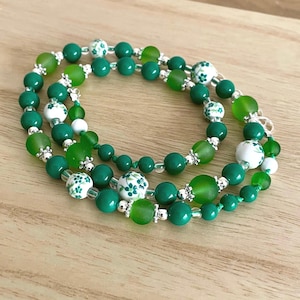May include: A green and white beaded necklace with floral patterns. The necklace is made of multiple strands of beads, including green glass beads, white ceramic beads with floral designs, and silver spacer beads.