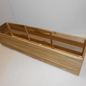 May include: A rectangular wooden planter box with a natural wood finish. The box has a long, narrow shape with vertical wooden slats forming the sides. Interior supports are visible. Ideal for outdoor or indoor plants.