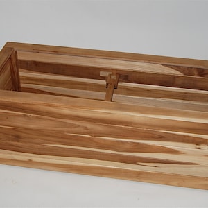 May include: A rectangular wooden planter box with a natural finish. The box is constructed from horizontal wooden planks and features a simple, open design. The interior has a central support. The planter is suitable for outdoor or indoor use.