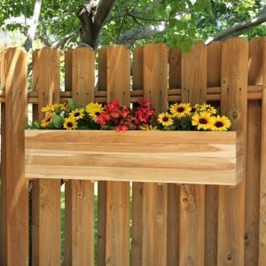 May include: A rectangular wooden planter box filled with yellow and red flowers, mounted on a wooden fence. The planter box is made of light-colored wood and is filled with a variety of colorful flowers.
