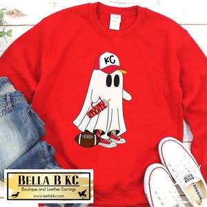 May include: A bright red crewneck sweatshirt featuring a graphic of a white ghost wearing a red and white baseball cap with the letters KC, red high-top sneakers, and holding a red patterned tumbler next to a brown football.