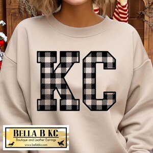 May include: A beige crewneck sweatshirt featuring the letters KC in a black and white buffalo plaid pattern. The garment has long sleeves and a round neckline. A rectangular label at the bottom displays the text BELLA B KC Boutique and Leather Earrings.