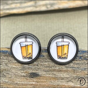 May include: A pair of stud earrings with a cartoon bubble tea design. The earrings have a black metal border and a white background. The bubble tea is orange with brown tapioca pearls.