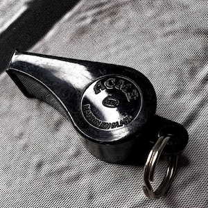 May include: A black whistle with a metal key ring. The whistle has the text "ACME" and "MADE IN ENGLAND" printed on it. The whistle is made of a shiny, smooth material.