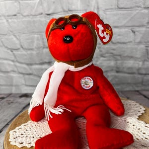 May include: A red plush bear wearing a brown aviator cap and goggles, a white scarf, and a patch that reads "100 Years of Flight." The bear is a collectible item, likely a Beanie Baby.