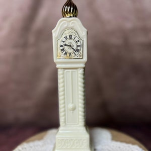May include: A cream-colored, miniature grandfather clock figurine with a gold-colored top. The clock face features black Roman numerals and gold accents. The figurine is displayed on a wooden surface with a lace doily.