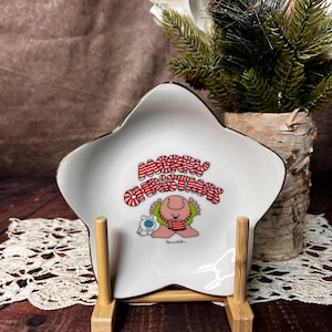 May include: A white star-shaped ceramic dish with a gold rim. The dish features a cartoon character with a red and white striped sweater and the words "Merry Christmas". The dish is on a wooden stand.