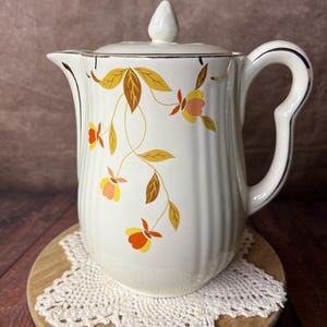 May include: A cream-colored ceramic coffee pot with a ribbed body and a matching lid. The exterior features a decorative pattern of stylized orange, yellow, and brown flowers with thin vines. The rim and handle are accented with a thin gold trim.