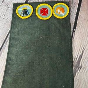 May include: A green fabric sash with three embroidered badges. The badges are yellow with a green centre and feature a railway track, a red cross, and a white shoe with a pumpkin.