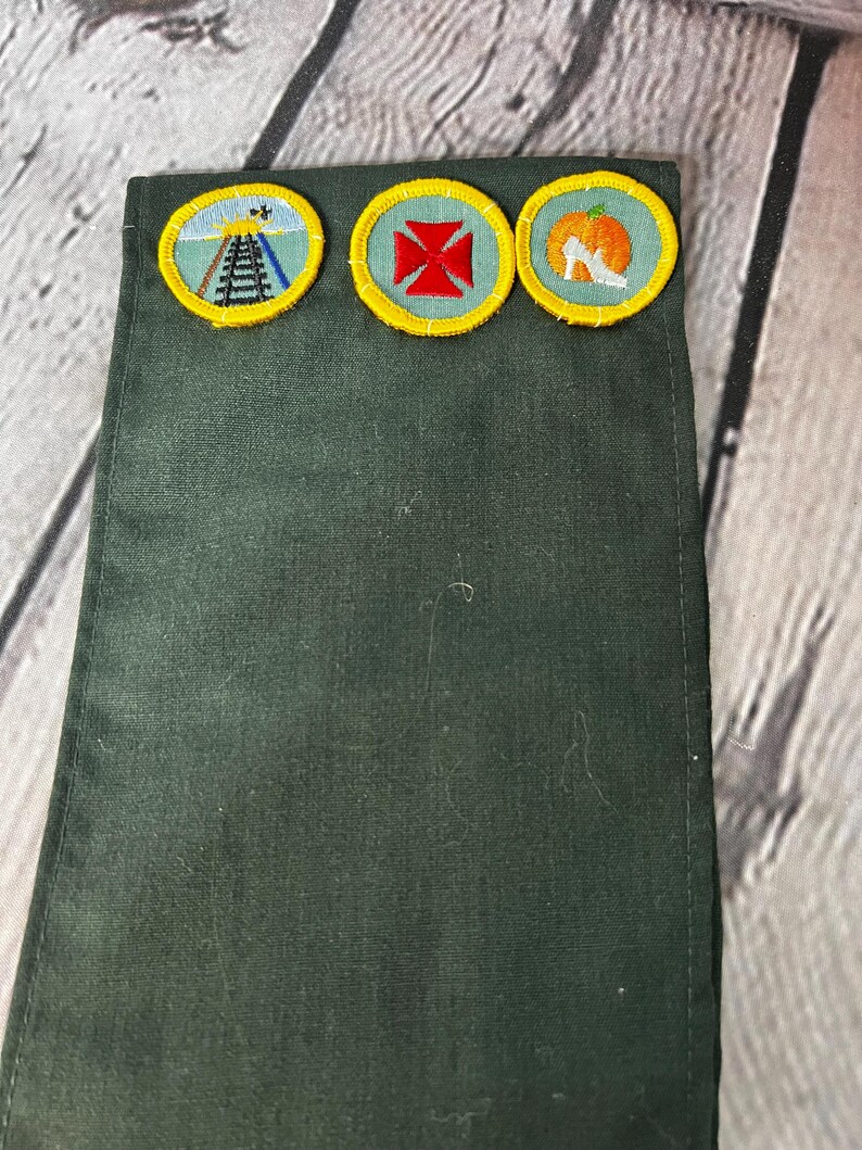 May include: A green fabric sash with three embroidered badges. The badges are yellow with a green centre and feature a railway track, a red cross, and a white shoe with a pumpkin.