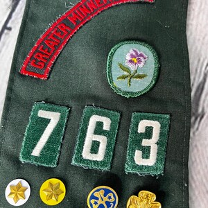 May include: Green Girl Guide sash with a red patch that says "GREATER MINNEAPOLIS", three green patches with white numbers "763", a purple flower patch, and several gold and blue badges.