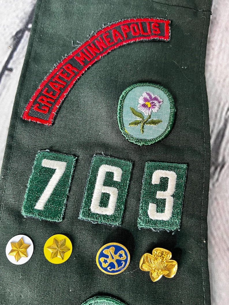 May include: Green Girl Guide sash with a red patch that says "GREATER MINNEAPOLIS", three green patches with white numbers "763", a purple flower patch, and several gold and blue badges.