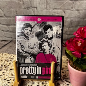 May include: DVD of the movie "Pretty in Pink" with a black and white image of the cast. The DVD is displayed on a small wooden stand. The title is in pink and white. A pink flower in a pink pot is in the background.