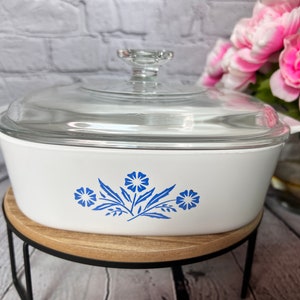 May include: White casserole dish with a glass lid and a blue floral design. The dish is sitting on a wooden stand.