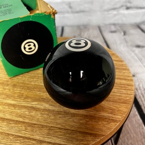 May include: A black 8-ball with a white number 8 on it. The ball is sitting on a wooden table.