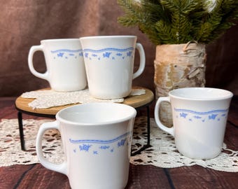 May include: Four white ceramic mugs with a blue floral design. The mugs have a handle and a straight, cylindrical shape. The mugs are arranged on a wooden surface and a lace doily. The mugs are a classic style.