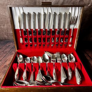May include: A vintage silverware set displayed in a wooden box with a red velvet interior. The set includes knives, forks, and spoons, all with ornate silver handles. The box is open, revealing the neatly arranged utensils.