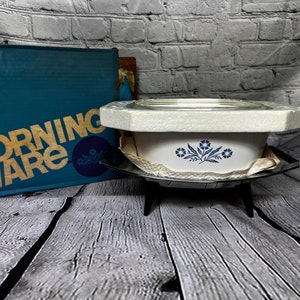 May include: A white CorningWare casserole dish with a blue floral design, in its original box. The dish is on a metal stand with three legs.
