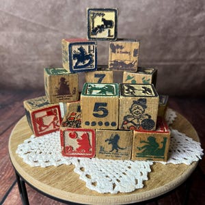May include: A collection of vintage wooden alphabet and number blocks stacked in a pyramid. The blocks feature various illustrations including a clown, a bear, a rocking horse, and the number 5, printed in black, red, and green on natural wood.