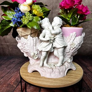May include: A white ceramic planter with two cherubs holding a bouquet of flowers. The planter is decorated with pink and white flowers and has a tree trunk design. The planter is sitting on a wooden stand with a black metal base.