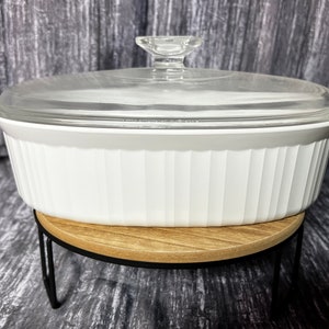 May include: White oval baking dish with a glass lid, sitting on a wooden and black metal stand.