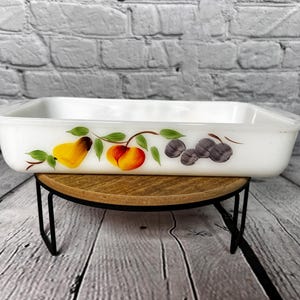 May include: White rectangular baking dish with a painted design of fruit and leaves. The dish is on a wooden stand with black metal legs.