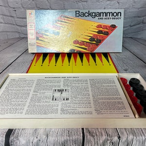 May include: A vintage Milton Bradley backgammon game set in a white box with a yellow and black game board and red and black playing pieces. The box lid features the game name "Backgammon and Acey-Deucy".