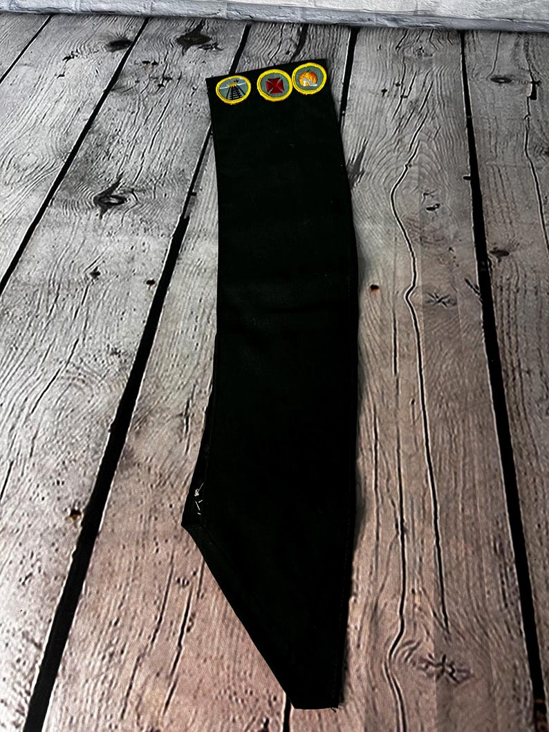 May include: A black fabric sash with three yellow and brown scout badges attached. The badges feature a railway crossing, a campfire, and a compass.