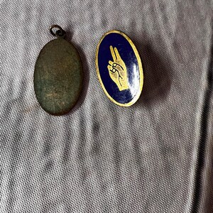 Op de afbeelding: Twee vintage metalen spelden. E&eacute;n is een ovale hanger met een lus aan de bovenkant. De andere is een ovale speld met een blauwe achtergrond en een gouden handgebaar. Het handgebaar toont de wijs- en middelvinger omhoog.