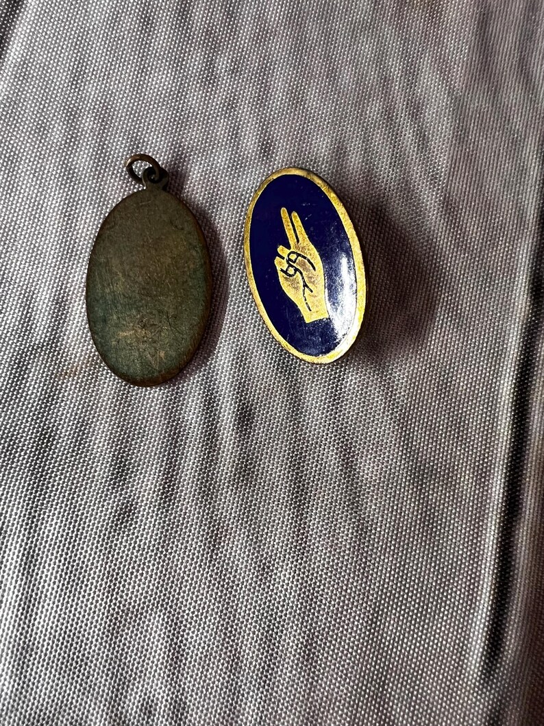 Op de afbeelding: Twee vintage metalen spelden. E&eacute;n is een ovale hanger met een lus aan de bovenkant. De andere is een ovale speld met een blauwe achtergrond en een gouden handgebaar. Het handgebaar toont de wijs- en middelvinger omhoog.